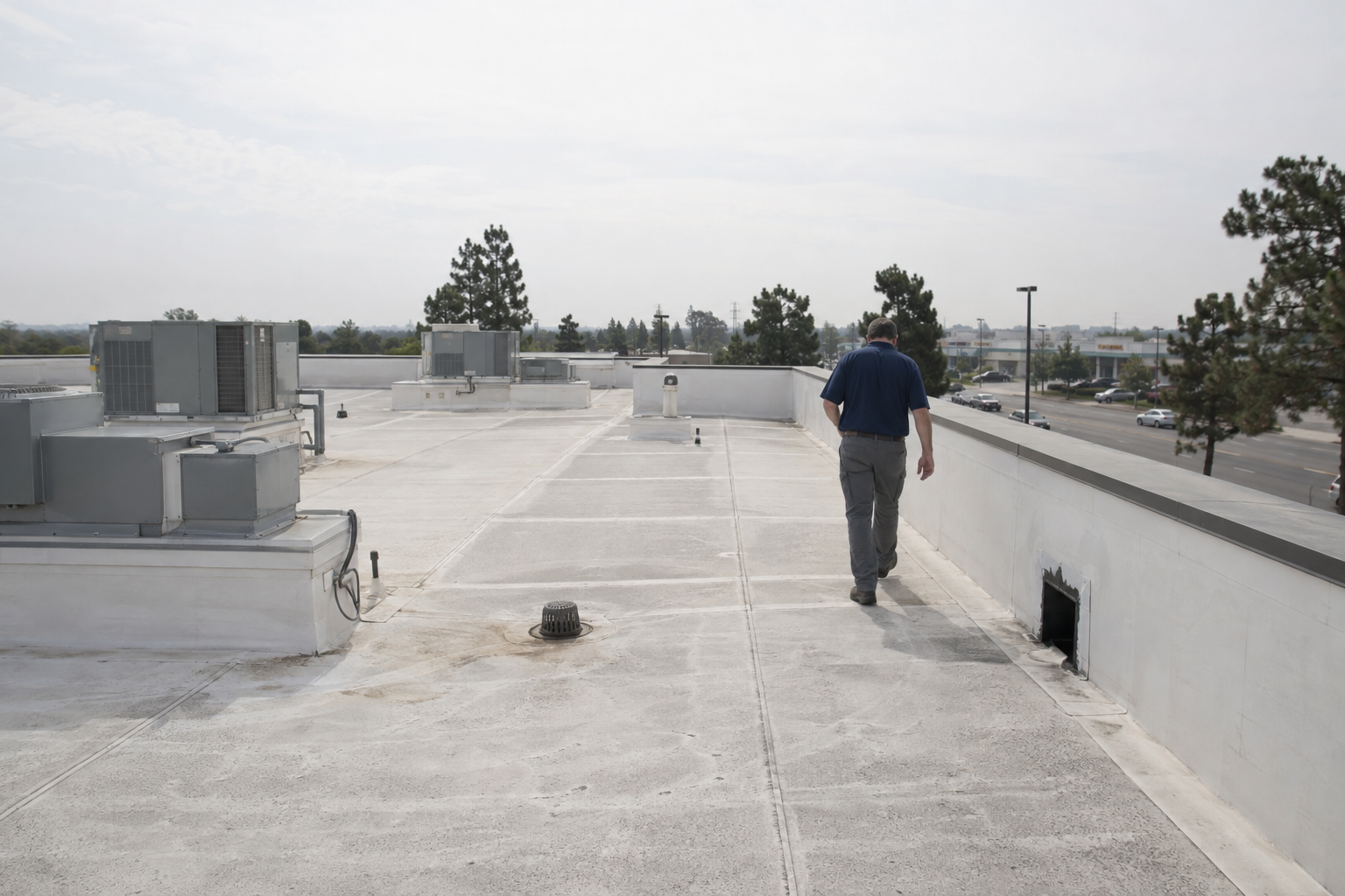 Roofer walking across a low-slope commercial roof toward a scupper drain past rooftop units