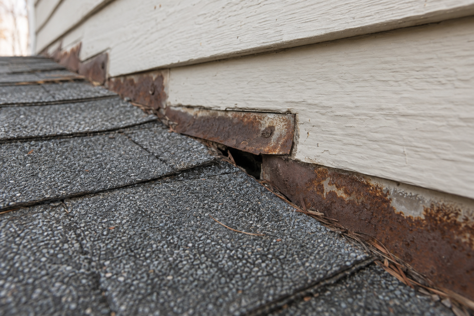 Corroded step flashing with an open gap where a shingle roof meets a painted siding wall